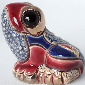 May include: A ceramic frog figurine with a red, blue, and gold glaze. The frog has large, black eyes and a textured surface.