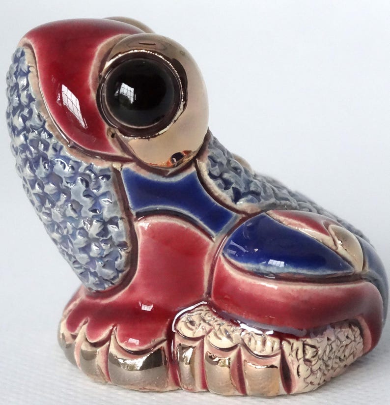 May include: A ceramic frog figurine with a red, blue, and gold glaze. The frog has large, black eyes and a textured surface.