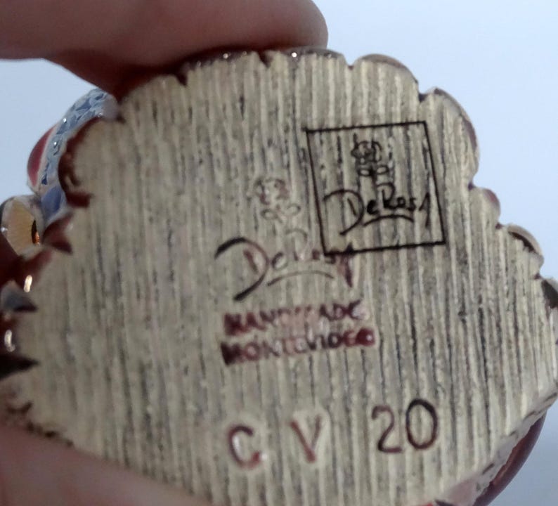 May include: Close-up of the bottom of a ceramic figurine. The figurine is signed "De Rossi" and "Handmade in Montevideo". The number "CV 20" is also visible.