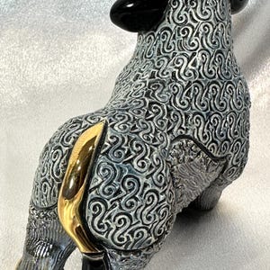 May include: A ceramic bull figurine featuring a textured grey and black swirling pattern across its body. The bull has polished gold-colored horns, a gold-colored tail, and gold-colored hooves, with solid black ears and a black patch on its head.