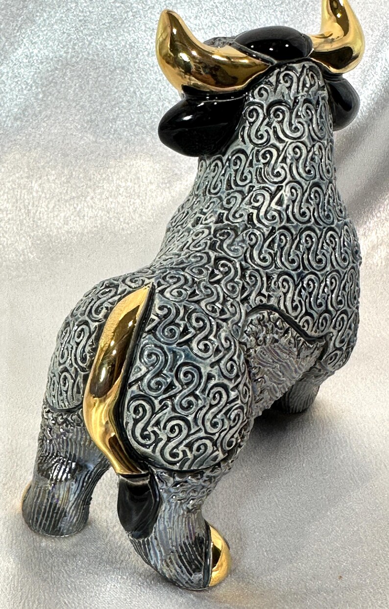 May include: A ceramic bull figurine featuring a textured grey and black swirling pattern across its body. The bull has polished gold-colored horns, a gold-colored tail, and gold-colored hooves, with solid black ears and a black patch on its head.