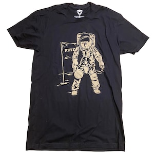 May include: Black t-shirt with a graphic of an astronaut playing basketball on the moon. The astronaut is wearing a spacesuit and holding a basketball. The text "Pete" is visible on a sign behind the astronaut.