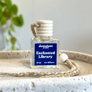May include: A clear glass car air freshener bottle with a wooden cap and a blue label that reads "Sunnsheen scents Enchanted Library 10 ml car diffuser". The bottle is suspended by a beige and gold rope.