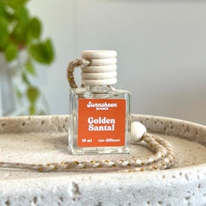 May include: A clear, square car air freshener bottle with a wooden cap and a woven cord. The label is orange with the text "Sunnsheen scents Golden Santal 10 ml car diffuser". The bottle is sitting on a light-colored, textured surface.
