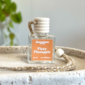 May include: A clear, square car air freshener bottle with a wooden cap and a braided cord. The label reads "Sunnsheen scents Fizzy Pineapple 10 ml car diffuser". The bottle is on a light-colored, textured surface.