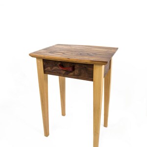 May include: A wooden end table with a single drawer and a red leather pull. The table has a dark wood top and light wood legs.