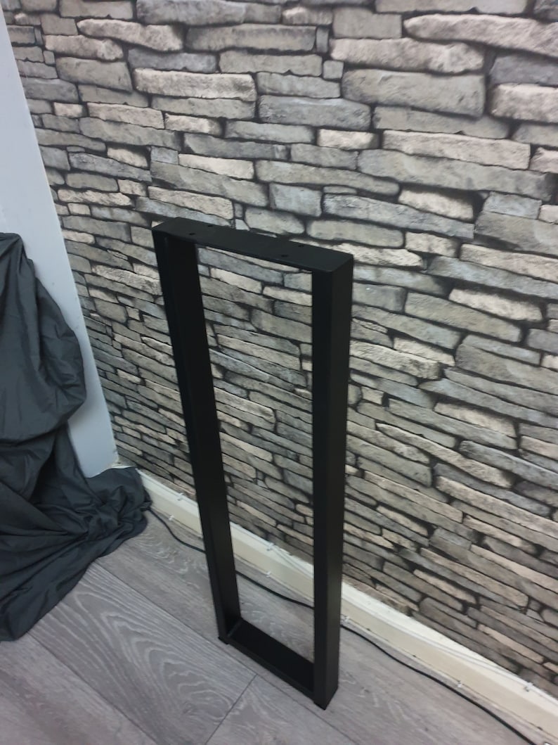 May include: Black metal table leg with a rectangular frame. The leg is standing on a wood floor.