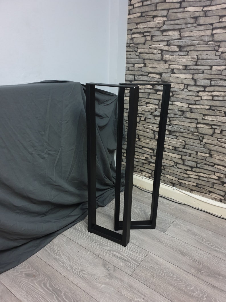 May include: Two black metal table legs, each with a rectangular shape and a square base. The legs are standing on a light gray wood floor.