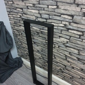 May include: A tall, rectangular black metal furniture leg frame with a hollow center. The frame features a flat top bar with pre-drilled circular holes for mounting, two long vertical side bars, and a solid horizontal base bar at the bottom.