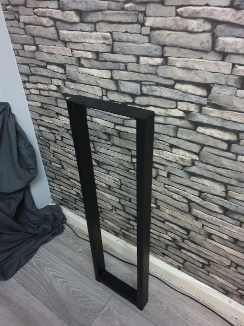 May include: A tall, rectangular black metal furniture leg frame with a hollow center. The frame features a flat top bar with pre-drilled circular holes for mounting, two long vertical side bars, and a solid horizontal base bar at the bottom.