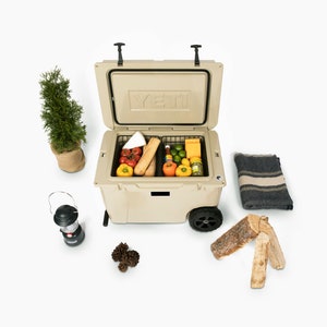 Puede incluir: Nevera rodante de color beige con la tapa abierta, que revela alimentos como tomates, pimientos y pan. Un peque&ntilde;o &aacute;rbol, una linterna, una manta y le&ntilde;a est&aacute;n dispuestos alrededor de la nevera, sugiriendo un picnic o reuni&oacute;n al aire libre.