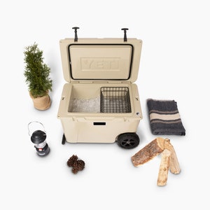 Puede incluir: Nevera beige con la tapa abierta, que revela hielo y una cesta de alambre. Un peque&ntilde;o &aacute;rbol de hoja perenne en una maceta de arpillera, una linterna, pi&ntilde;as, una manta y le&ntilde;a sugieren un tema de acampada.