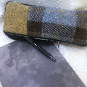 Może przedstawiać: Szary notes z srebrnym wzorem gwiazdy i czarnym długopisem leżącym na nim. Na notesie leży brązowa, niebieska i żółta pikowana kosmetyczka z zamkiem błyskawicznym.