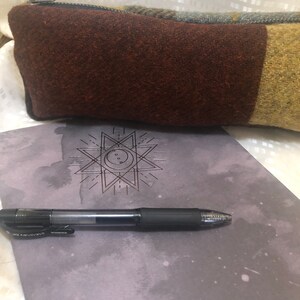 May include: A brown and green wool pencil case with a zipper closure. The pencil case is lying on a grey notebook with a geometric star design and a black pen.