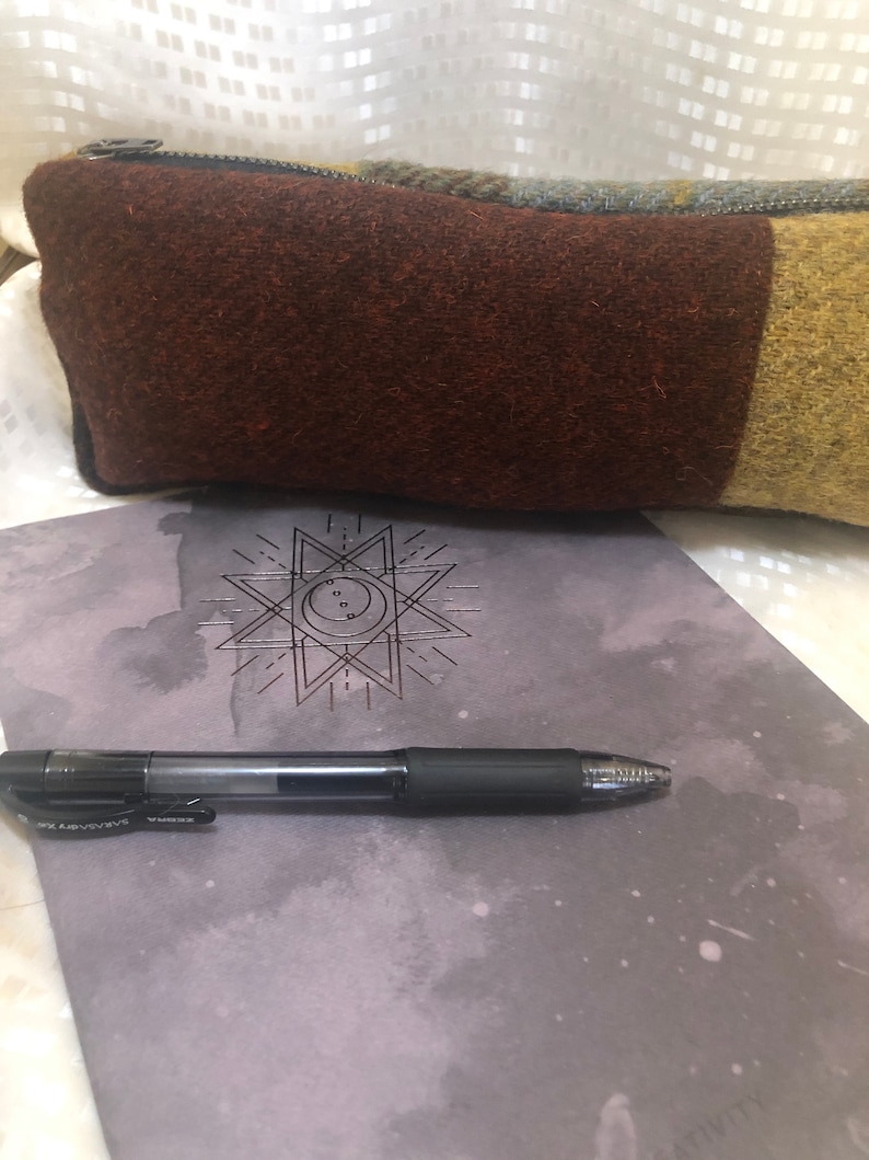May include: A brown and green wool pencil case with a zipper closure. The pencil case is lying on a grey notebook with a geometric star design and a black pen.