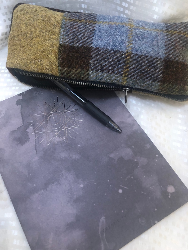 May include: A grey notebook with a silver star design and a black pen lying on top of it. A brown, blue and yellow plaid zipped pouch is lying on top of the notebook.