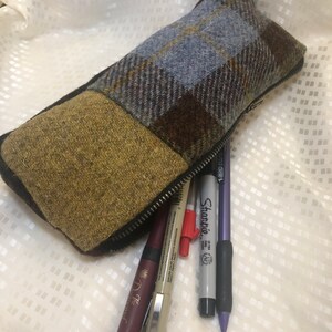 May include: A brown, blue and yellow plaid pencil case with a zipper closure. The case is open and filled with pens and pencils, including a purple Sharpie.