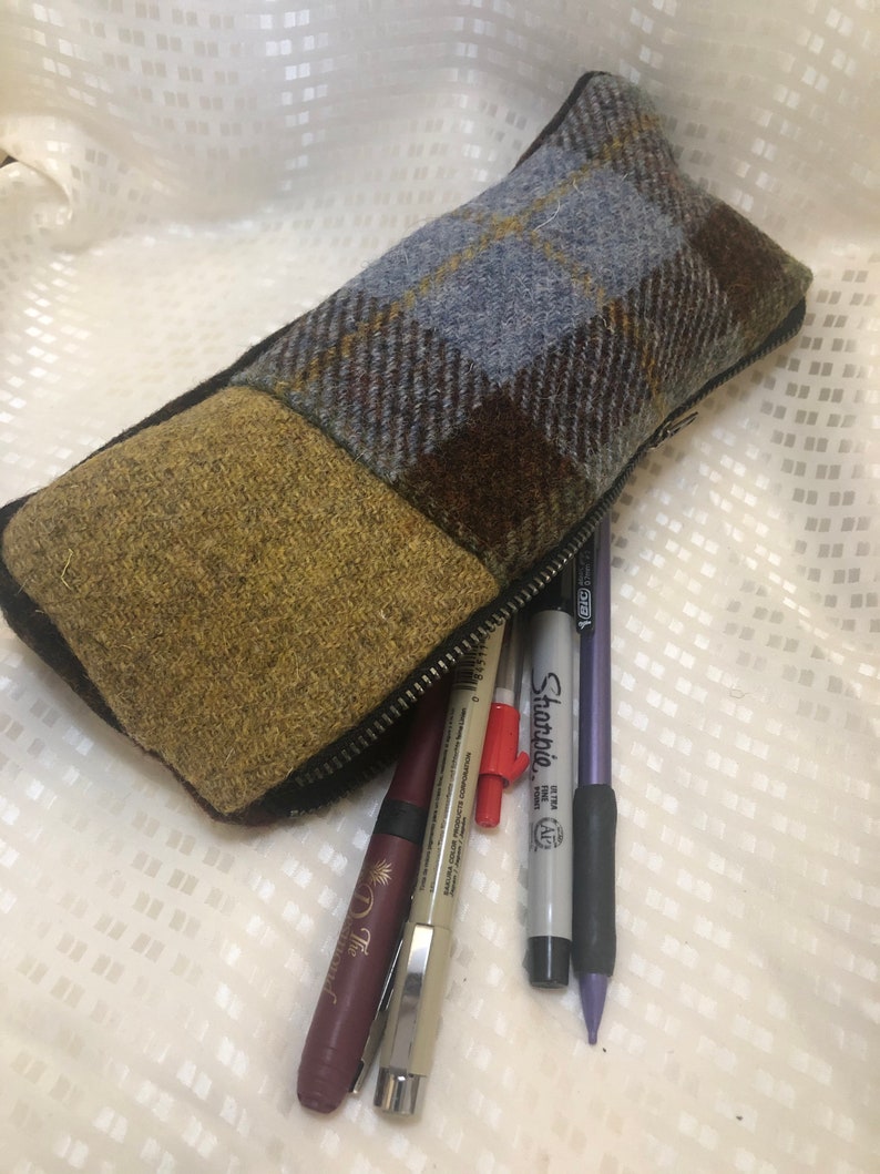 May include: A brown, blue and yellow plaid pencil case with a zipper closure. The case is open and filled with pens and pencils, including a purple Sharpie.