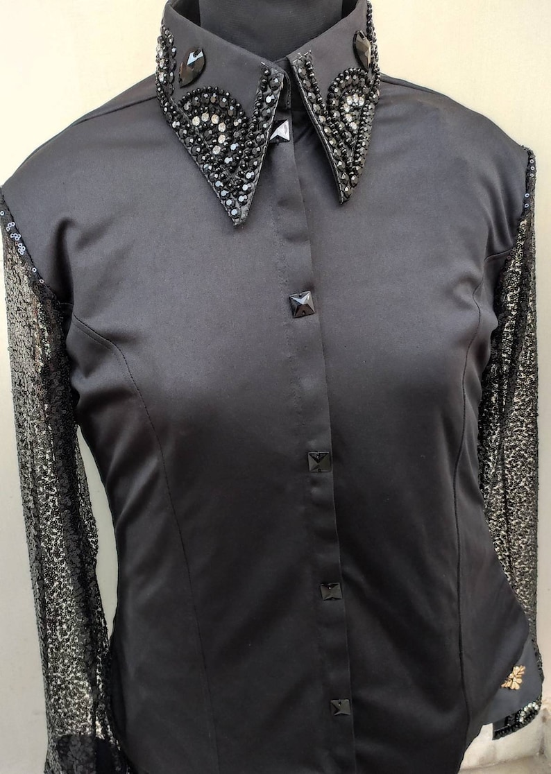 Puede incluir: Una camisa negra de botones con un cuello grande adornado. El cuello est&aacute; decorado con diamantes de imitaci&oacute;n y lentejuelas negras. La camisa tiene mangas largas con una superposici&oacute;n de encaje negro.