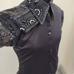 Puede incluir: Una camisa negra de botones con cuello y mangas de lentejuelas. El cuello est&aacute; adornado con pedrer&iacute;a negra y plateada en un dise&ntilde;o floral.