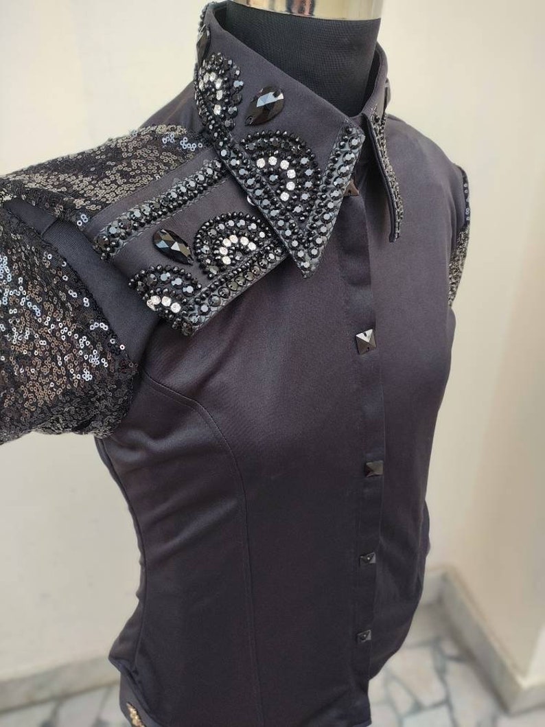 Puede incluir: Una camisa negra de botones con cuello y mangas de lentejuelas. El cuello est&aacute; adornado con pedrer&iacute;a negra y plateada en un dise&ntilde;o floral.