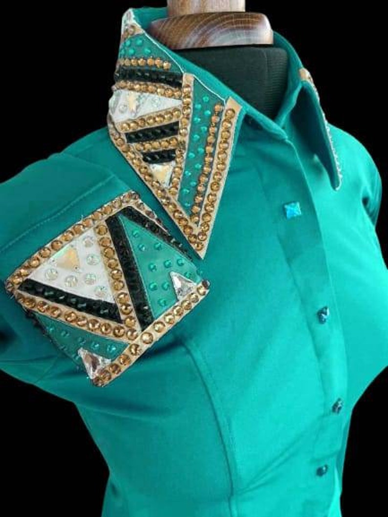 May include: Teal long-sleeve shirt with a gold, black, and green geometric design on the collar and one sleeve. The design features rhinestones and a white fabric accent.
