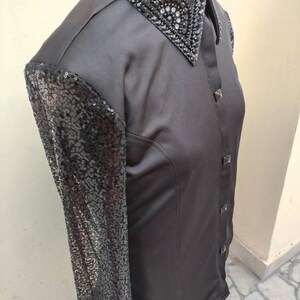 Puede incluir: Una camisa negra de manga larga con una manga de lentejuelas y un cuello negro adornado. El cuello est&aacute; decorado con pedrer&iacute;a y cuentas negras.