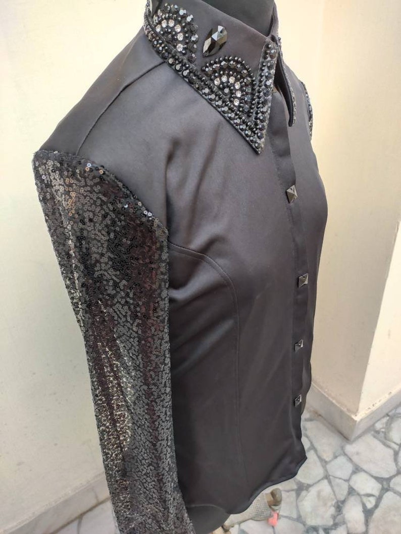 Puede incluir: Una camisa negra de manga larga con una manga de lentejuelas y un cuello negro adornado. El cuello est&aacute; decorado con pedrer&iacute;a y cuentas negras.