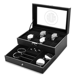 Puede incluir: Caja de reloj de cuero negro con tapa de vidrio y un caj&oacute;n. La caja tiene un monograma en la parte superior y contiene varios relojes, anillos, pulseras y gafas de sol.