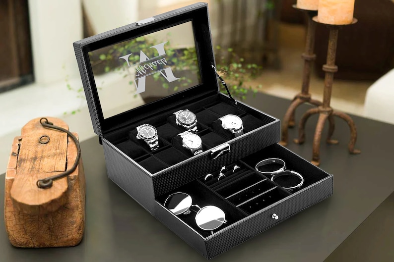 Puede incluir: Una caja de reloj de cuero negro con tapa de vidrio y un monograma personalizado. La caja tiene dos niveles, el nivel superior tiene ranuras para relojes y el nivel inferior tiene compartimentos para joyas y gafas de sol.