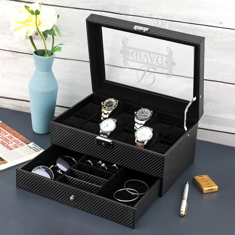 Puede incluir: Caja de reloj de cuero negro con tapa de vidrio y un caj&oacute;n. La caja tiene un patr&oacute;n de fibra de carbono y est&aacute; personalizada con el nombre "DAVID". La caja tiene ranuras para varios relojes y un caj&oacute;n para guardar otros accesorios.