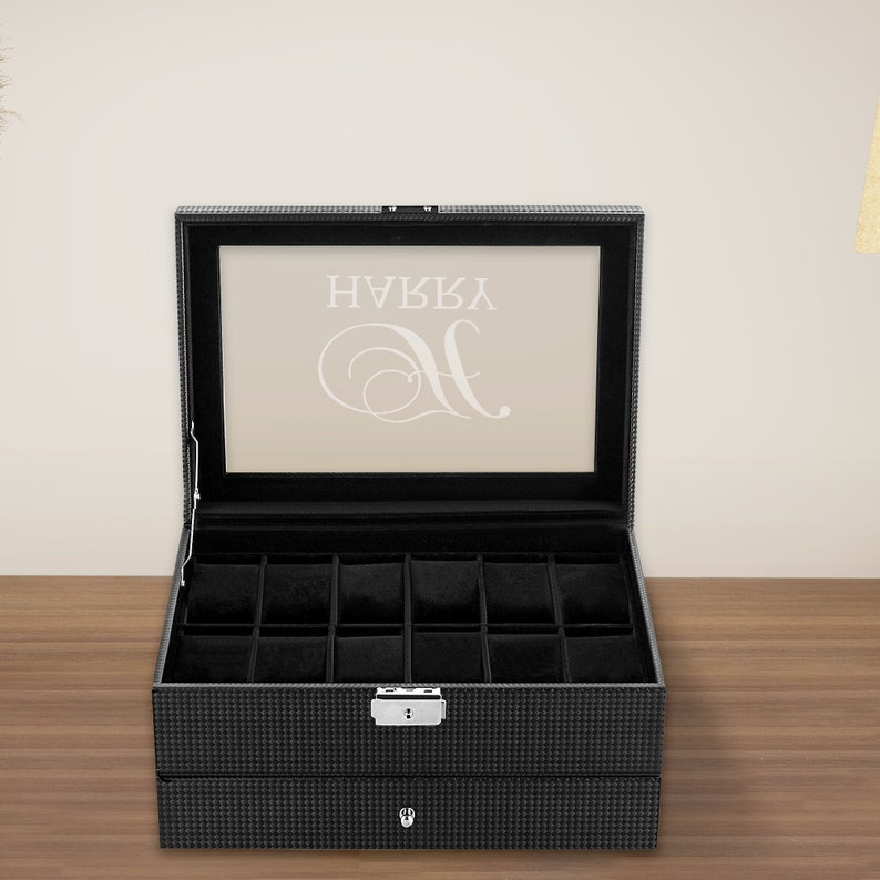 Puede incluir: Caja de reloj de cuero negro con tapa de vidrio y un caj&oacute;n. La caja tiene 12 compartimentos para relojes y una cerradura en la parte delantera. La tapa de vidrio tiene el texto "NAVY" y un monograma "H" en blanco.