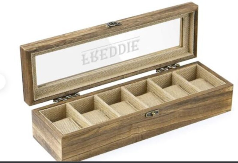 May include: A wooden watch box with a glass top and six compartments lined with brown fabric. The box is engraved with the name "LKEDDIE".