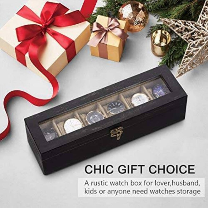 May include: A black wooden watch box with a glass top and six compartments for storing watches. The box has a gold latch and is sitting on a white surface.