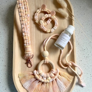 May include: A set of handmade macrame items in a peach and cream colour scheme. The set includes a keychain, a necklace with a wooden bead and a tassel, and a pair of earrings. The items are displayed on a wooden tray with a bottle of essential oil labelled "Aromist Stress Relief".