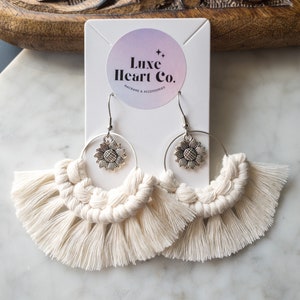 May include: Cream macrame earrings with a fan design. Each earring has a silver sunflower charm and a circular hoop. The earrings are displayed on a white card with the text "Luxe Heart Co."