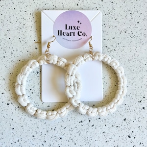 May include: A pair of white macrame hoop earrings with gold hooks. The earrings are displayed on a white card with the text "Luxe Heart Co. Macrame & Accessories".