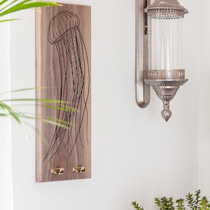 Puede incluir: Un colgador de llaves de pared de madera con un dibujo de línea negra de una medusa. El colgador de llaves tiene dos ganchos de latón.