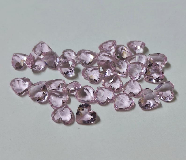 May include: A collection of heart-shaped, faceted gemstones in a soft pink hue. The gems are scattered, catching the light and displaying multiple facets. These could be used for jewelry making or other craft projects.