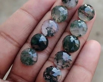 May include: A collection of faceted, round gemstones with moss-like inclusions. The stones display a mix of clear, green, and dark hues. The gemstones are held in a hand, showcasing their polished surfaces and unique patterns. These stones are suitable for jewelry making.