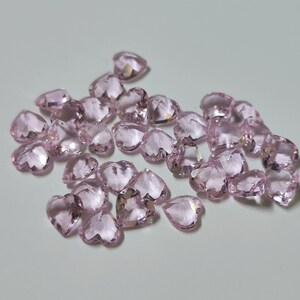 May include: A collection of faceted, heart-shaped gemstones in a soft pink hue. The gemstones are scattered across a light-colored surface, catching the light and displaying multiple facets. The overall effect is one of delicate beauty and sparkle.