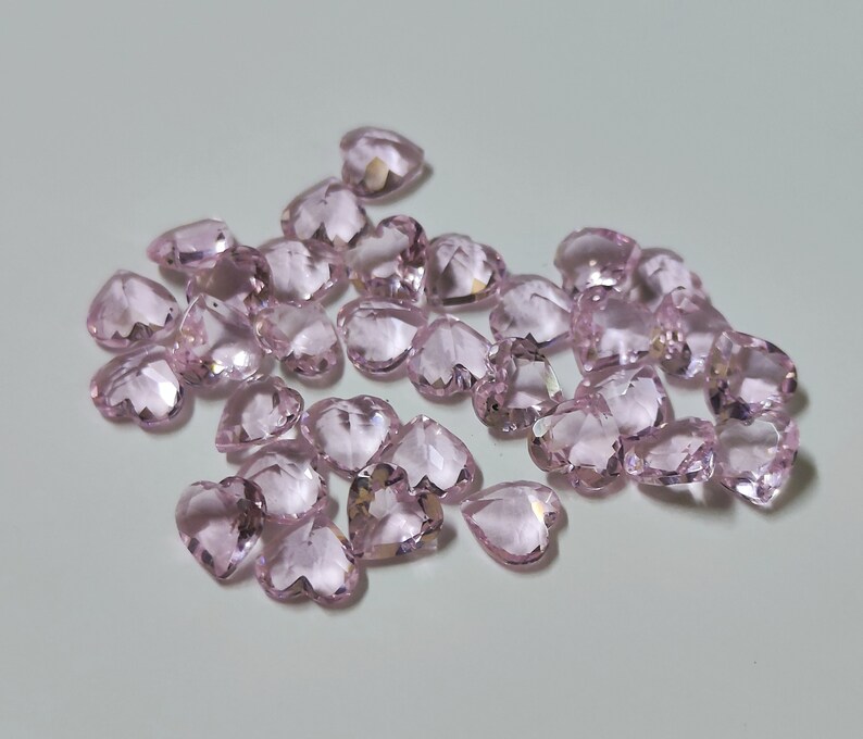 May include: A collection of faceted, heart-shaped gemstones in a soft pink hue. The gemstones are scattered across a light-colored surface, catching the light and displaying multiple facets. The overall effect is one of delicate beauty and sparkle.
