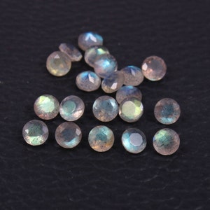 May include: A collection of faceted, round labradorite gemstones. The stones display a mix of gray, blue, and green iridescence. The gemstones are scattered on a dark surface, showcasing their polished surfaces.
