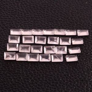May include: A selection of rectangular, pale pink gemstones displayed on a dark background. The gemstones are translucent and polished, reflecting light. The stones vary in size, indicating they are cut from a larger piece. Measurements are in cm.