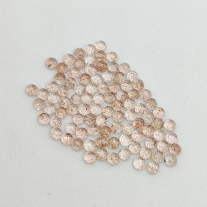 May include: A collection of small, round, faceted gemstones. The gems are primarily clear with reddish-brown inclusions, scattered across a white surface. The overall appearance suggests a natural, earthy aesthetic, suitable for jewelry making.