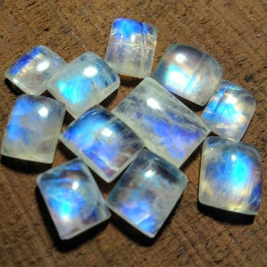 May include: A collection of rectangular moonstone cabochons with rounded corners. The stones display a milky white base with iridescent blue and yellow flashes. The gems are arranged on a dark brown wooden surface, creating a contrast that highlights their colors.