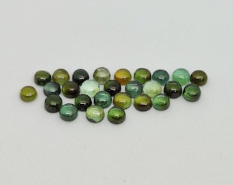 Puede incluir: Una colección de gemas redondas y pulidas en varios tonos de verde, desde oliva claro hasta verde bosque oscuro. Las piedras, talladas en cabujón, están dispuestas sobre una superficie blanca, mostrando sus superficies lisas y abovedadas y sus variaciones de color.