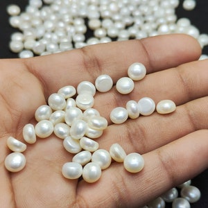 May include: A collection of lustrous, white, half-drilled pearls. The pearls are round or slightly flattened, with a smooth, pearlescent surface. Some pearls are scattered in the background. The pearls are ideal for jewelry making.