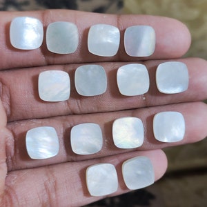 May include: A collection of twelve square, iridescent mother-of-pearl cabochons. Each cabochon has rounded corners and a pearlescent sheen, reflecting light with subtle variations in color. The cabochons are arranged on a neutral background.