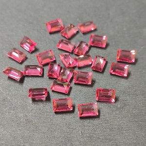 Puede incluir: Una colección de gemas rosas rectangulares y facetadas dispersas sobre una superficie gris oscuro. Las gemas tienen una apariencia vibrante y translúcida, con bordes afilados y un acabado pulido. Las gemas son probablemente para la fabricación de joyas.